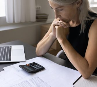 A woman rests her elbows on a gray desk while reviewing printed data with a laptop and a black calculator within reach.