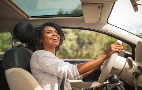 A smiling young Black woman is driving her car with her sunroof open. She's looking up at her rear-view mirror.