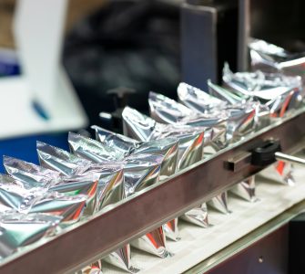 A machine holds several shiny, silver plastic pouches lined up in a row and secured by a metal arm bar.