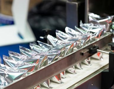 A machine holds several shiny, silver plastic pouches lined up in a row and secured by a metal arm bar.