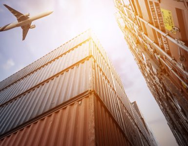 A low-angle shot of towering red and blue cargo containers. There is an airplane flying overhead in the sky.