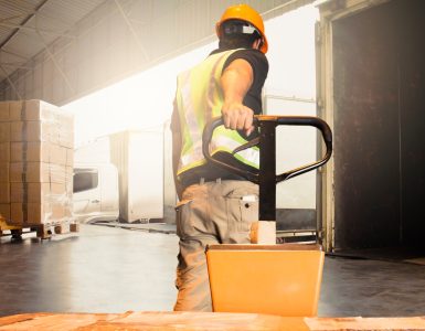 A man pulling a pallet jack toward a docking station at a warehouse. There are boxes wrapped in plastic on the equipment.