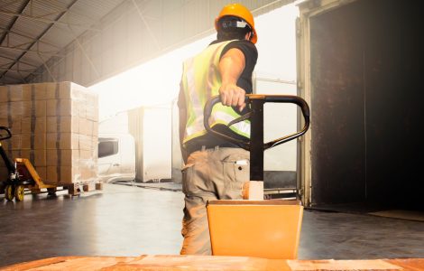 A man pulling a pallet jack toward a docking station at a warehouse. There are boxes wrapped in plastic on the equipment.