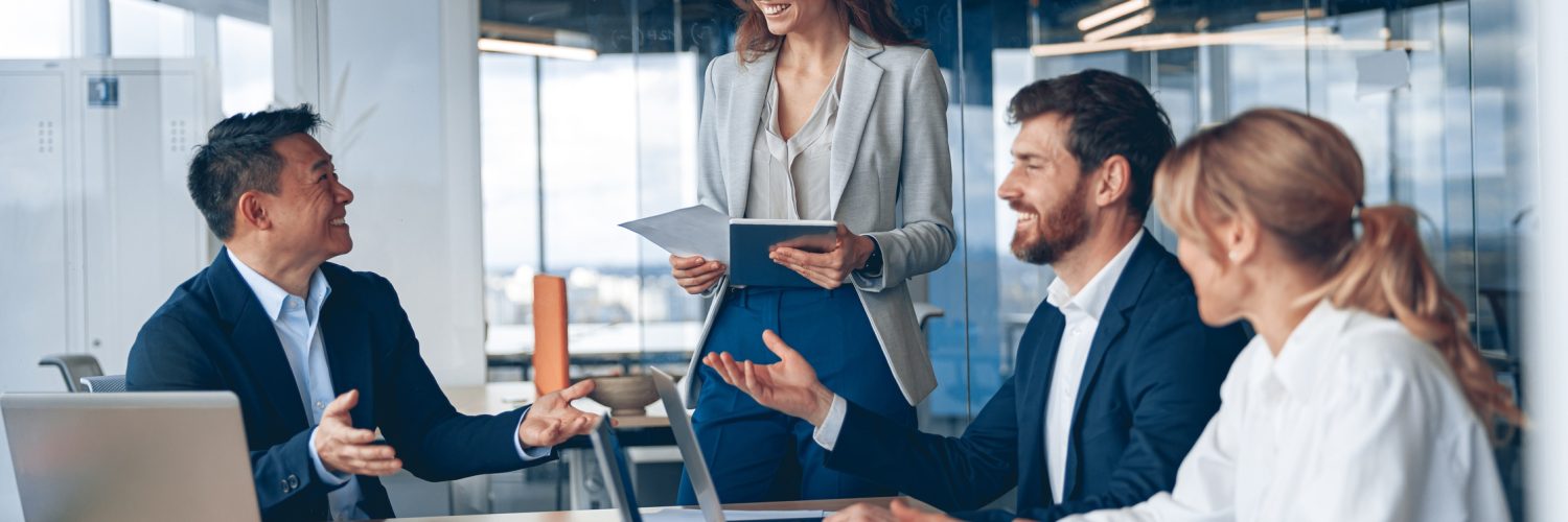 A female business professional stands in the conference room, with others sat at the table. She's presenting information.