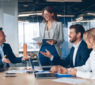 A female business professional stands in the conference room, with others sat at the table. She's presenting information.