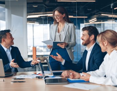 A female business professional stands in the conference room, with others sat at the table. She's presenting information.