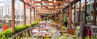 An outdoor dining patio features wooden beams with hanging string lights, wooden tables, and colorful metal chairs.