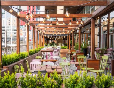 An outdoor dining patio features wooden beams with hanging string lights, wooden tables, and colorful metal chairs.