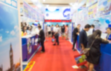 A blurred view of a crowd standing on a red carpet, surrounded by blue pop-up booths for a trade show.
