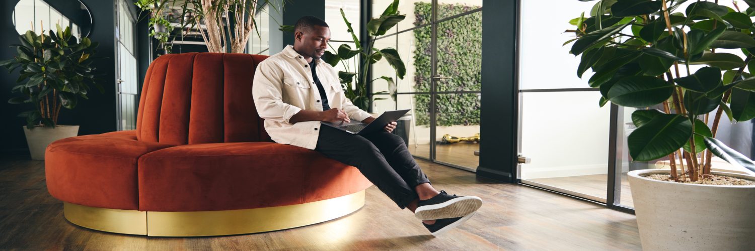 A man wearing a white shirt sits on an orange, circular sofa while browsing on a laptop, surrounded by potted plants.