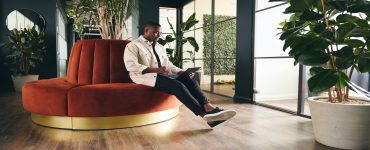 A man wearing a white shirt sits on an orange, circular sofa while browsing on a laptop, surrounded by potted plants.