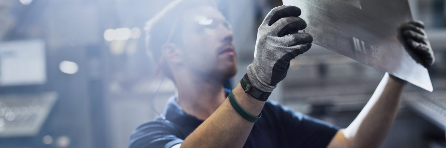 A young man wearing safety goggles and gloves holds a large sheet of metal with rectangular cutouts at a factory.