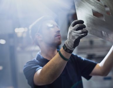 A young man wearing safety goggles and gloves holds a large sheet of metal with rectangular cutouts at a factory.