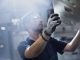 A young man wearing safety goggles and gloves holds a large sheet of metal with rectangular cutouts at a factory.