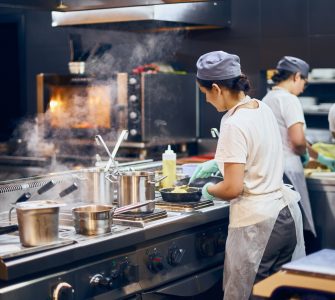 A team of cooks in the kitchen of a restaurant. They wear white shirts, aprons, and caps while working at different stations.