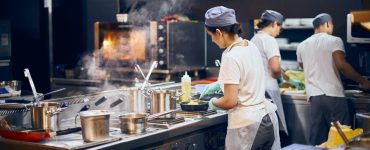 A team of cooks in the kitchen of a restaurant. They wear white shirts, aprons, and caps while working at different stations.
