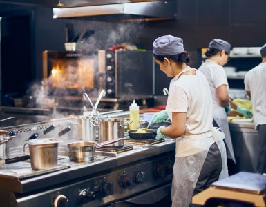 A team of cooks in the kitchen of a restaurant. They wear white shirts, aprons, and caps while working at different stations.