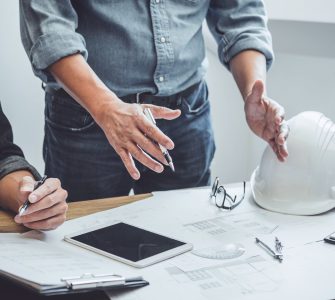 A table with construction company members standing around to discuss the blueprints. One person talks with their hands.