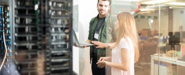 Two people stand in a server room separated from an office space by glass walls. One of them is holding a laptop.