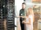 Two people stand in a server room separated from an office space by glass walls. One of them is holding a laptop.