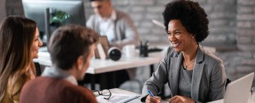 A woman in a gray blazer smiling while sitting across a desk from a man and woman. On the desk are stacks of paper and a laptop.