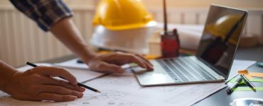 A person draws on the blueprint with one hand and uses a laptop on the other. Behind is a stack of hard hats.