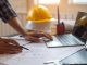 A person draws on the blueprint with one hand and uses a laptop on the other. Behind is a stack of hard hats.