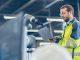 An engineer wearing a high-vis uniform uses a laptop while standing next to a robotic arm in a factory setting.