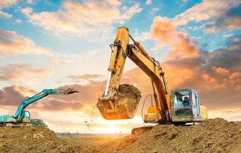 A yellow and light blue excavator working on a construction site. Behind them is the setting sun in a cloudy sky.