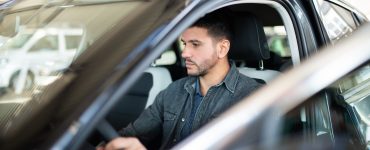 A man with a look of indifference on his face is sitting inside the driver’s seat of a car with the door open.