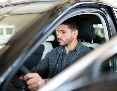 A man with a look of indifference on his face is sitting inside the driver’s seat of a car with the door open.