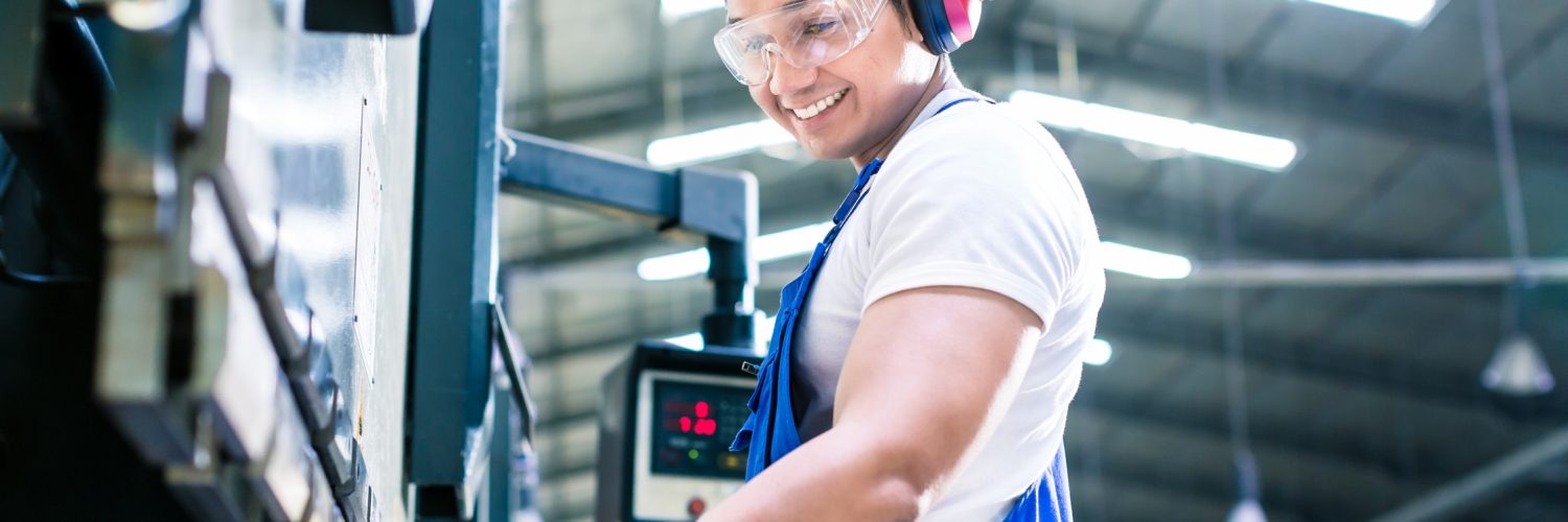 A male industrial worker wearing safety glasses and gloves is inspecting a manufactured piece in a factory.