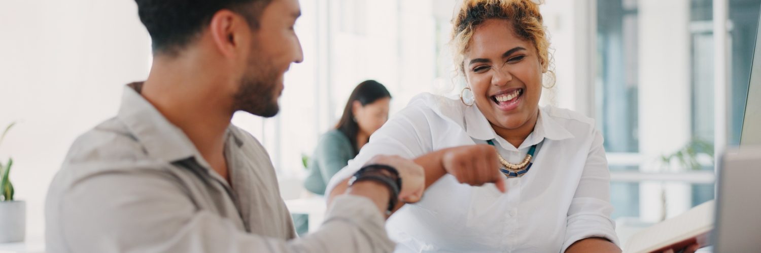 A man and a smiling woman fist-bump at a desk with a laptop and charts, while she holds an open notebook.