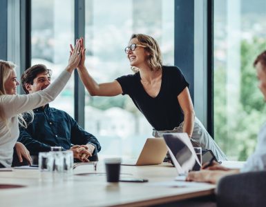 A woman standing at the head of a conference room table is high-fiving a woman sitting at the table. Two men sit at the table.