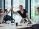 A woman standing at the head of a conference room table is high-fiving a woman sitting at the table. Two men sit at the table.