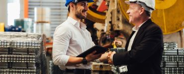 Engineer and manager wearing safety helmets shaking hands in manufacturing plant with equipment behind.