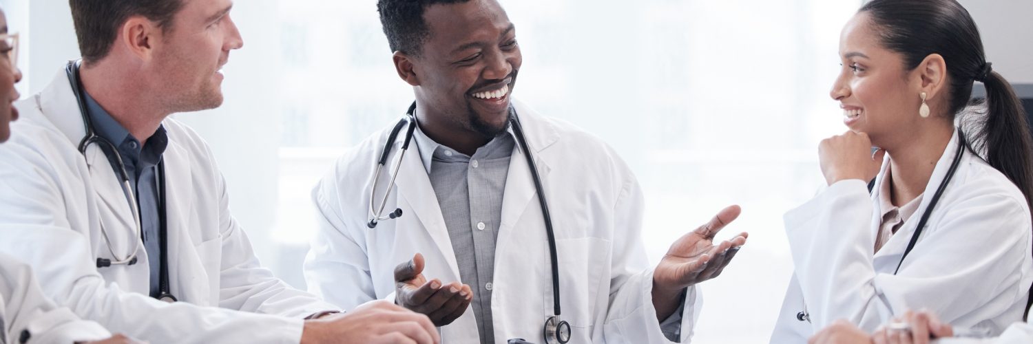 A group of doctors and healthcare staff sit around a table scattered with documents in a hospital meeting room.