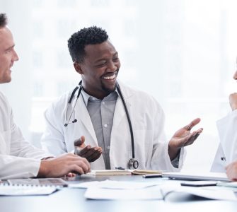 A group of doctors and healthcare staff sit around a table scattered with documents in a hospital meeting room.