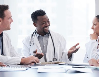 A group of doctors and healthcare staff sit around a table scattered with documents in a hospital meeting room.