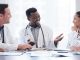 A group of doctors and healthcare staff sit around a table scattered with documents in a hospital meeting room.