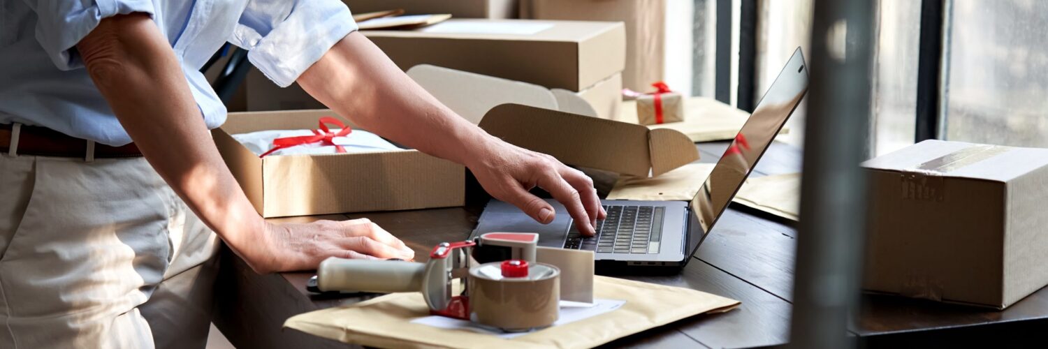 A person packaging orders at a workspace with boxes, tape, and a laptop, preparing items for shipping in a retail setting.