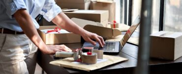 A person packaging orders at a workspace with boxes, tape, and a laptop, preparing items for shipping in a retail setting.