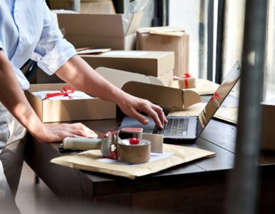 A person packaging orders at a workspace with boxes, tape, and a laptop, preparing items for shipping in a retail setting.