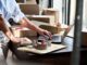 A person packaging orders at a workspace with boxes, tape, and a laptop, preparing items for shipping in a retail setting.