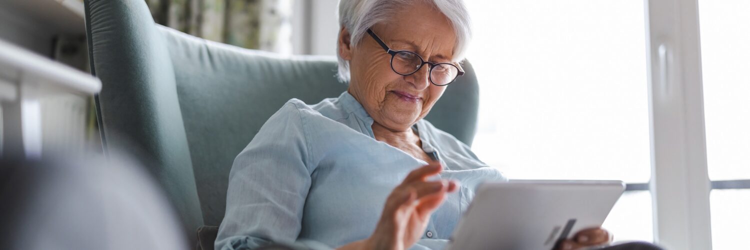 An older woman sitting in a blue chair inside with a tablet in her hands. There is a bright window behind her.