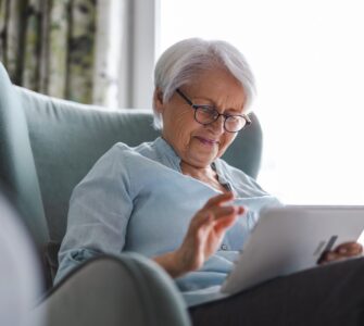 An older woman sitting in a blue chair inside with a tablet in her hands. There is a bright window behind her.