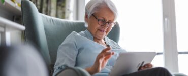 An older woman sitting in a blue chair inside with a tablet in her hands. There is a bright window behind her.