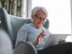 An older woman sitting in a blue chair inside with a tablet in her hands. There is a bright window behind her.