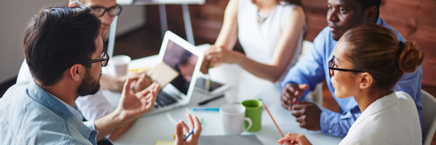 Multiple coworkers sit at a desk in an office while talking, with one man gesturing during the discussion.