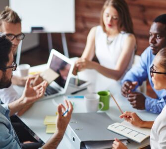 Multiple coworkers sit at a desk in an office while talking, with one man gesturing during the discussion.